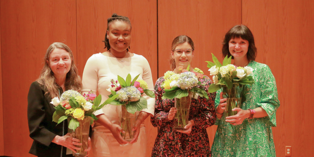 This photo is of four nicely dressed women standing shoulder to shoulder holding vases of flowers, accepting their Women of the Year awards