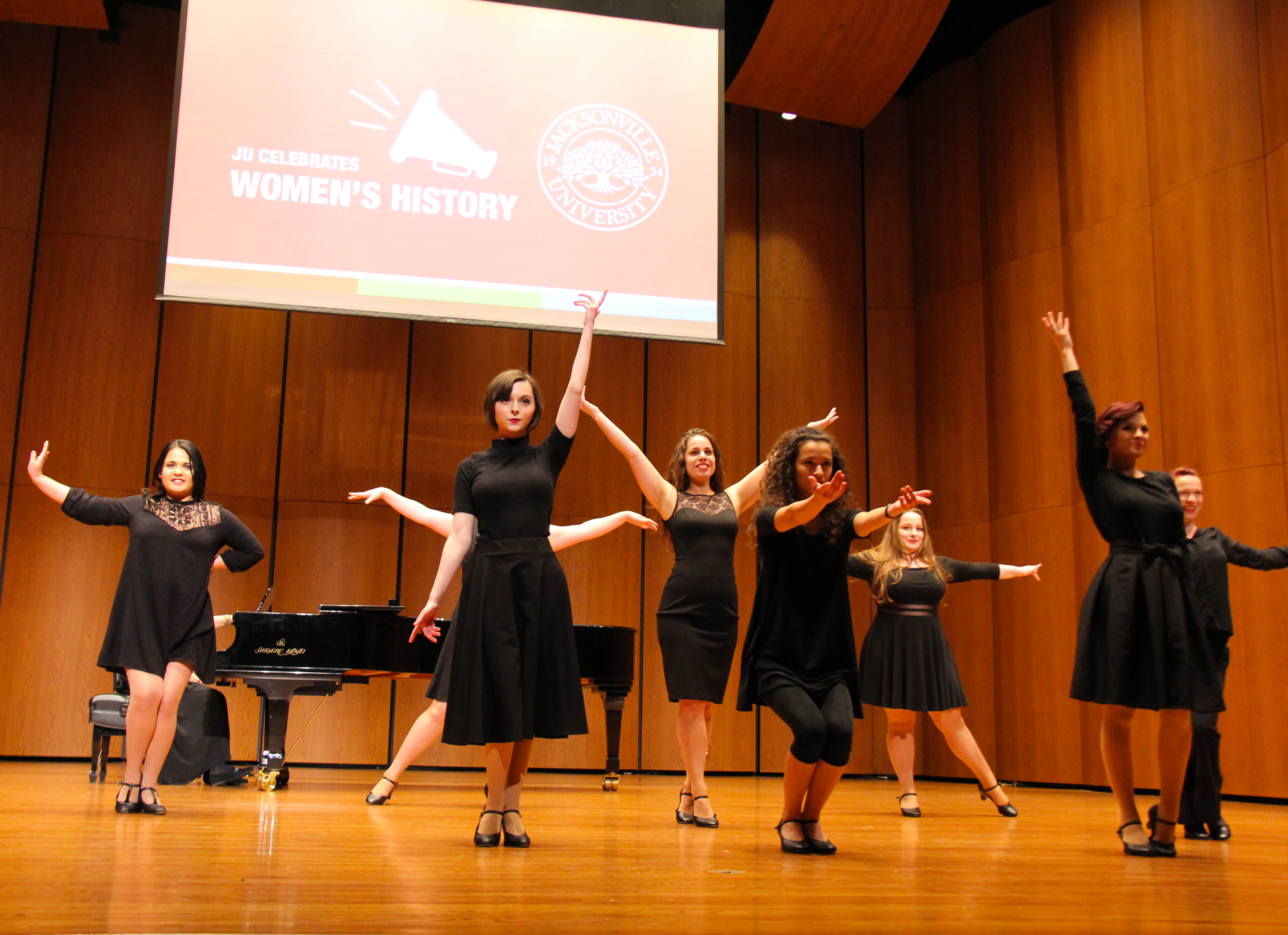 A female vocal ensemble in front of a Women's History Month sign, singing emotively