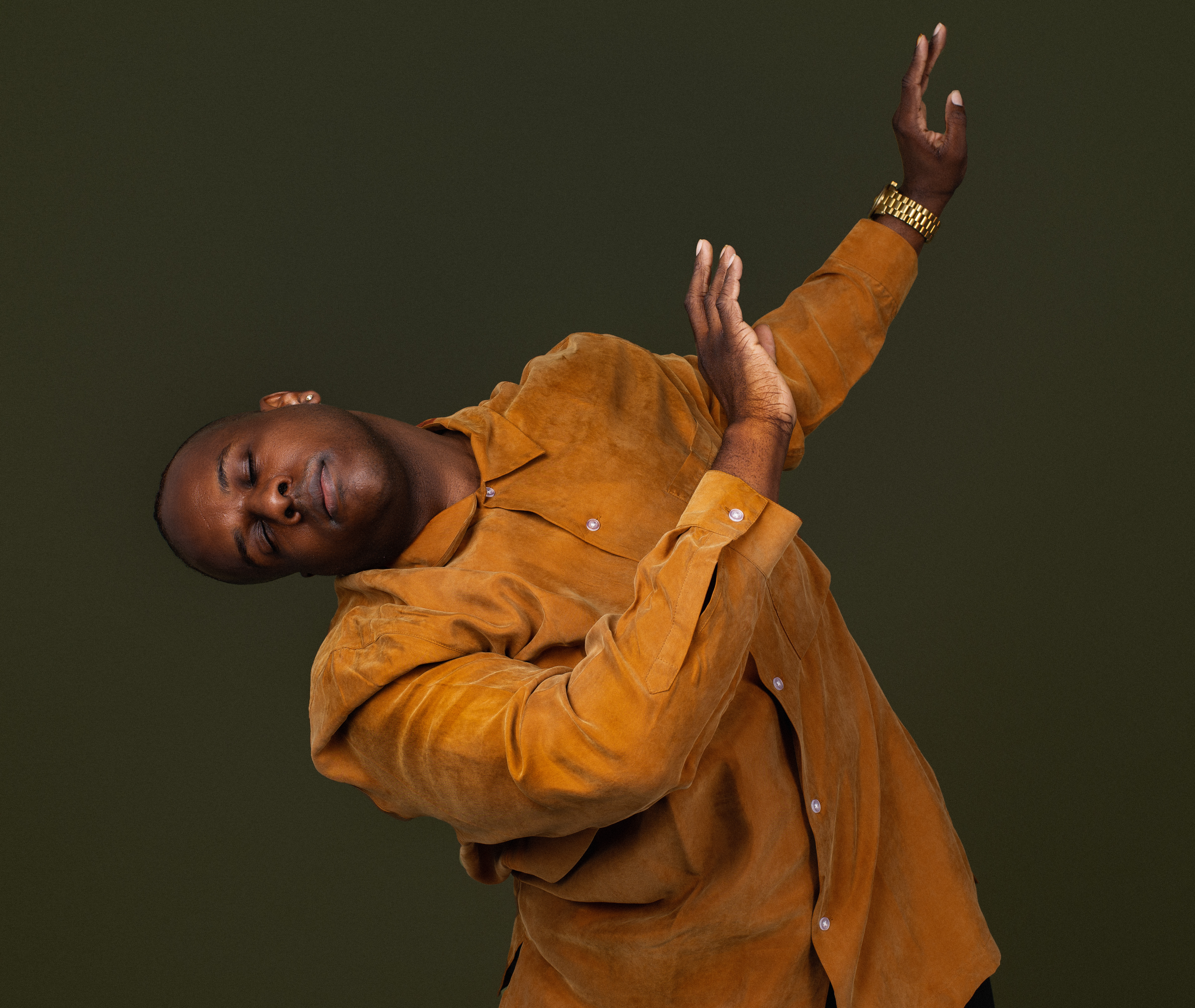 A Black choreographer and dancer is wearing a burnt orange tunic, leaning to the left with his body and gesturing to the right of the frame of the picture