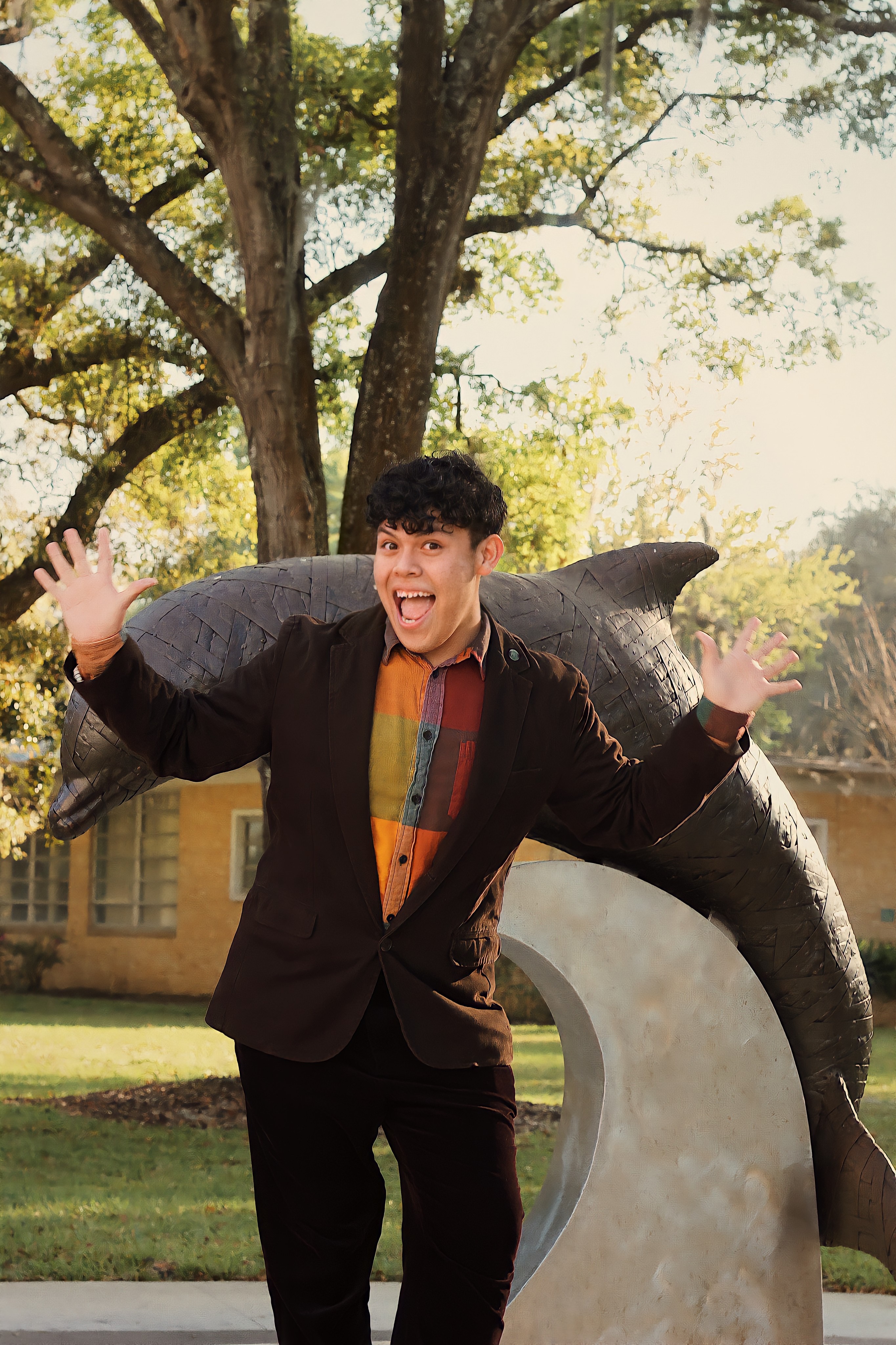 Male student in a colorful shirt and professional clothes standing near dolphin statues smiling with his hands raised