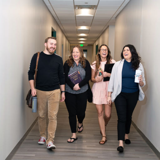 four students smiling and holding books and laptops while walking