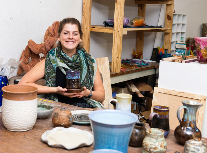 Student in the ceramics studio surrounded by ceramic and glass artwork.