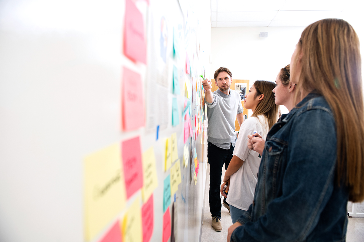 Students and professor engaged in a design thinking exercise using stick notes and collaborating on a writeboard wall.