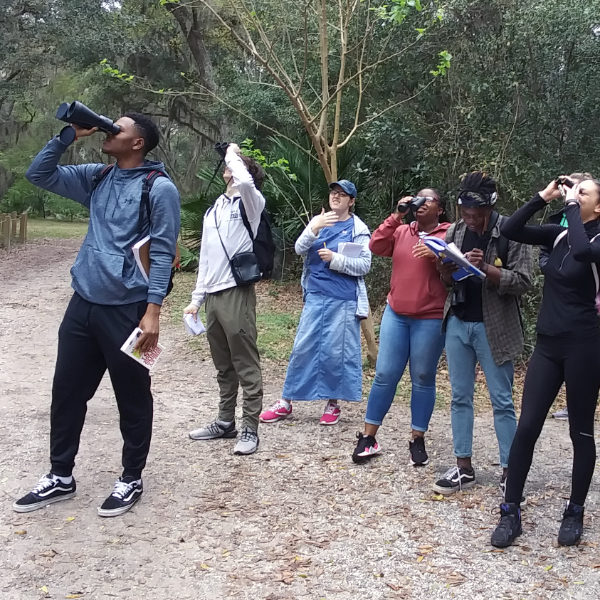A group of six student outdoors near trees looking up at the sky through binoculars.