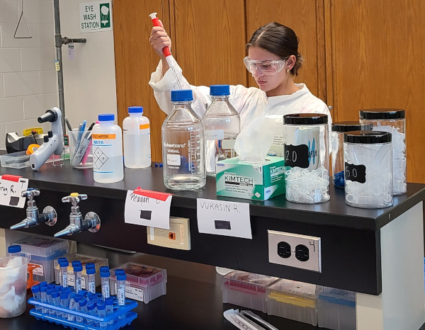 A young woman in a lab coat and safety goggles uses a red pipette at a lab bench with bottles, containers, and scientific equipment.