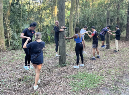 A photo of students on a wood trail holding hands around trees.