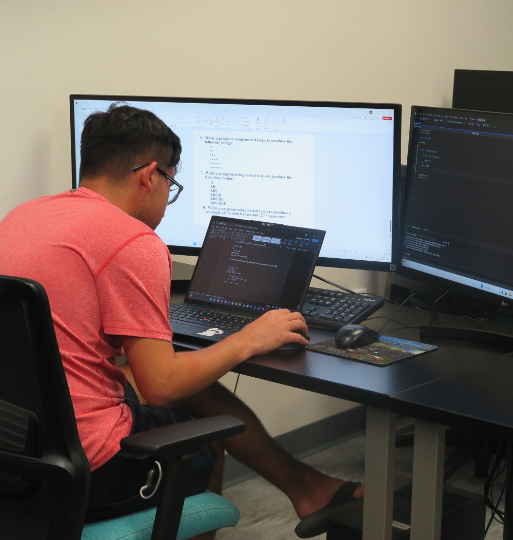 Student siting in front of a laptop and monitors working on sofware coding.