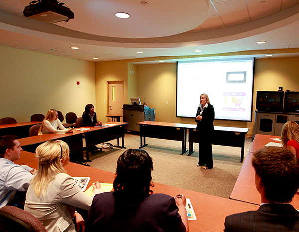 An adult woman speaks to a room full of adult students at tables arranged in a U shape around the room.