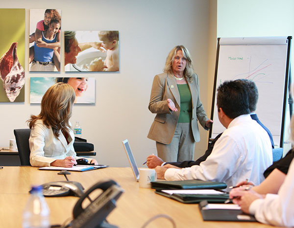 An adult woman presenting information from a chart to people gathered around a conference table.