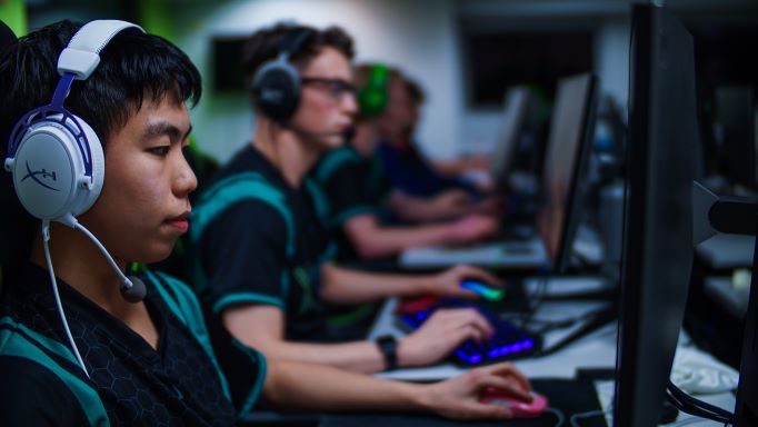 A male undergraduate student is facing a gaming monitor and wearing a white headset that has a microphone mouthpiece. The computer room is full of colors such as green and purple.