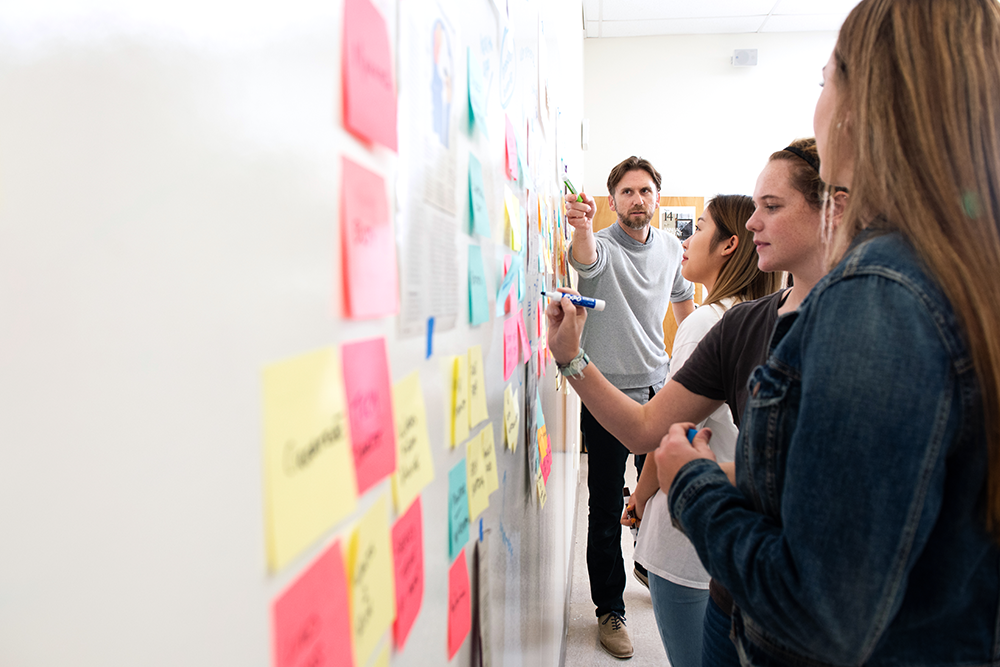 A male faculty member is standing on the far right of the picture and pointing at a wall full of colorful sticky notes, engaging with the onlooking students.
