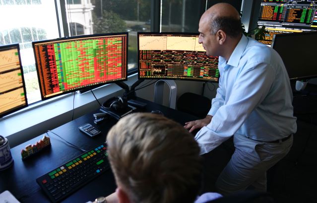 A nicely dressed male faculty member is working in the FinTech lab. He is standing beside a student and is in front of several dark monitors that have multicolored text on the screen.