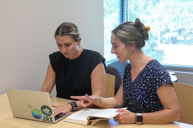 Two female graduate students talking and gesturing to one another while both looking a laptop screen..