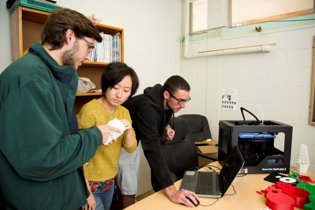 Three students are collaborating on a business project. One student is gesturing to the table in front of them which has a laptop on it, a printer, and a mix of red and green engineering equipment.