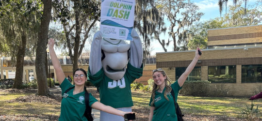 Dunk'n the mascot dolphin with two students lift up the Dolphin Dash flyer.