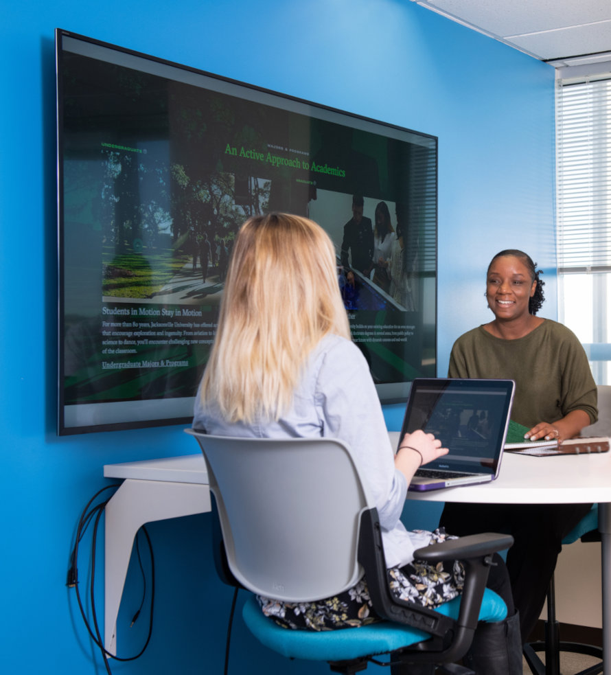 Two women seating at a desk with a laptop between them.