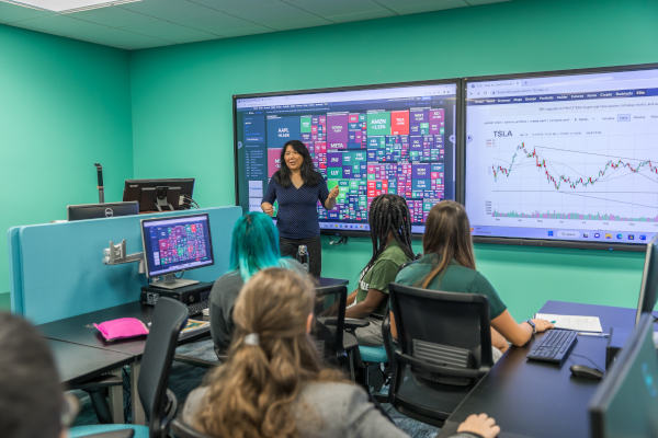 A professor standing near interactive screens presenting financial and market data to students in a modern classroom.