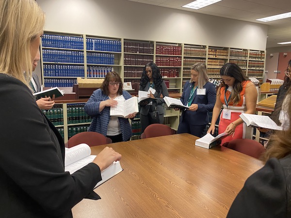 A law librarian standing at a table with a group of law students, showing them a section of a book she is holding up.
