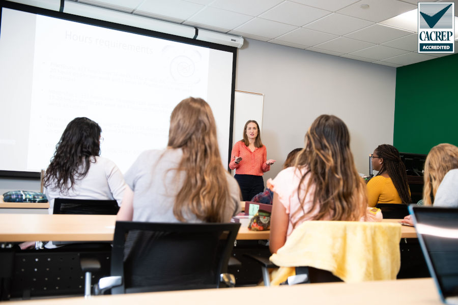 A teacher standing in front of a white board in a traditional classroom with students surrounding them.