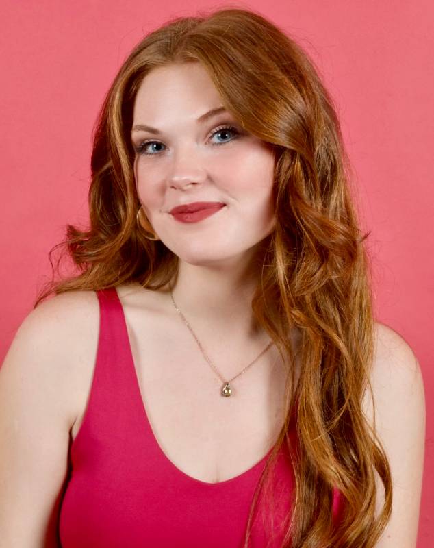 Profile photo of a red headed woman against a bubble gum pink backdrop, smiling in a pink tank top