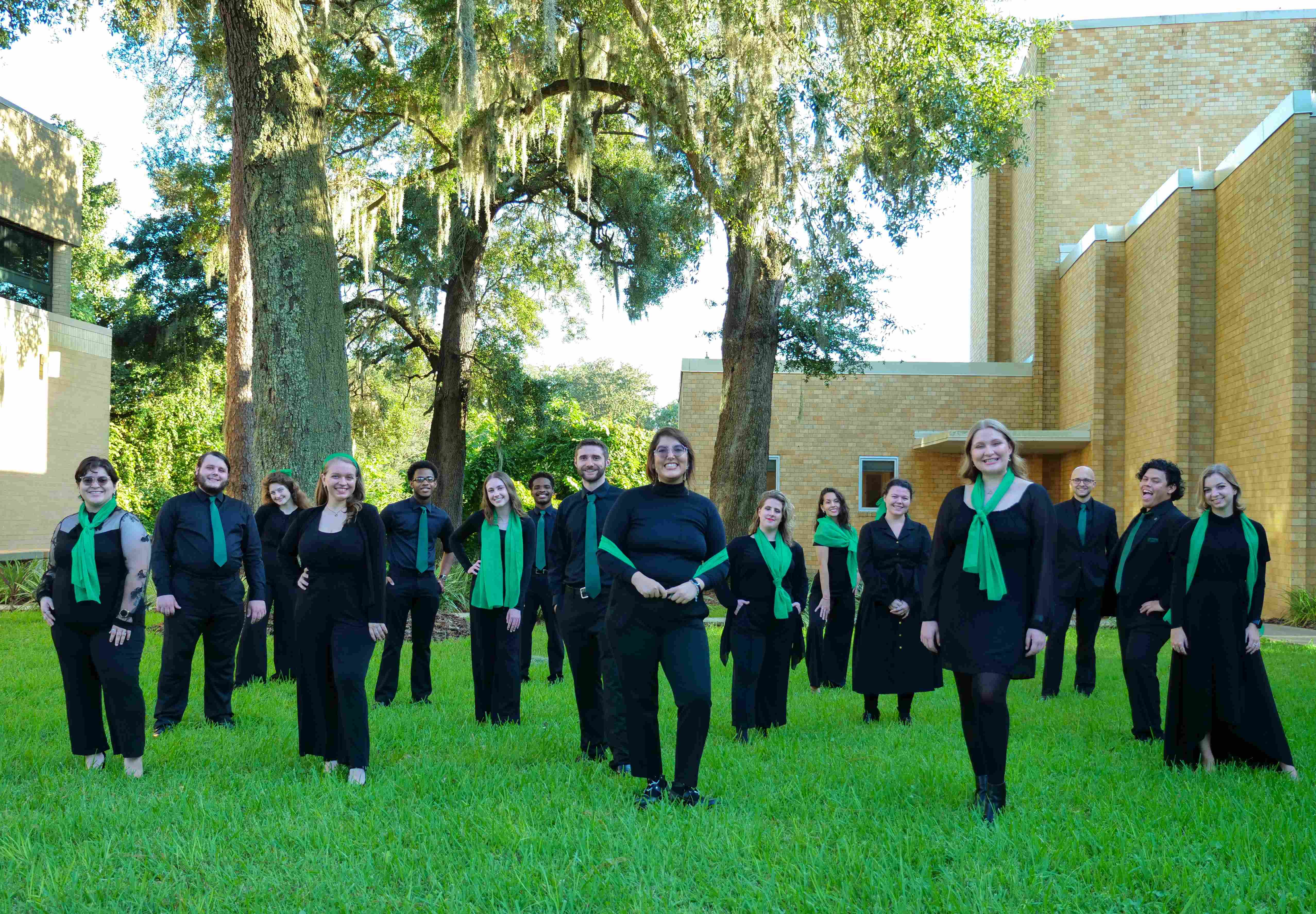 A large group of students wearing green scarves and ties over their "Concert black" attire are standing and smiling outside underneath the large oak trees characteristic of Jacksonville University's campus.   