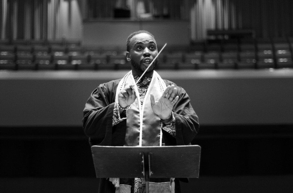 A black and white photo of a Black male student conductor wearing graduation regalia conducting an ensemble.