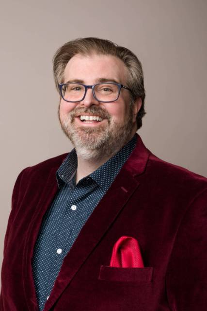 A man with short dark hair is smiling, wearing a velvet red suit, blue glasses, a blue shirt, and a red pocket square