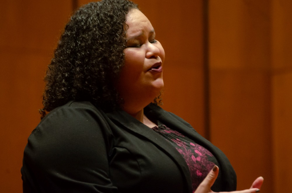 female student with dark curly hair in a dark suit and dress singing emotively 