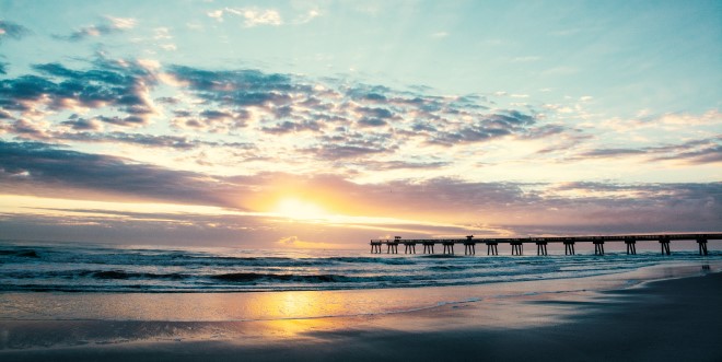 a picture of a beautiful sunrise over the beach next to the Jacksonville pier. The sky has blues, purples, and golds.