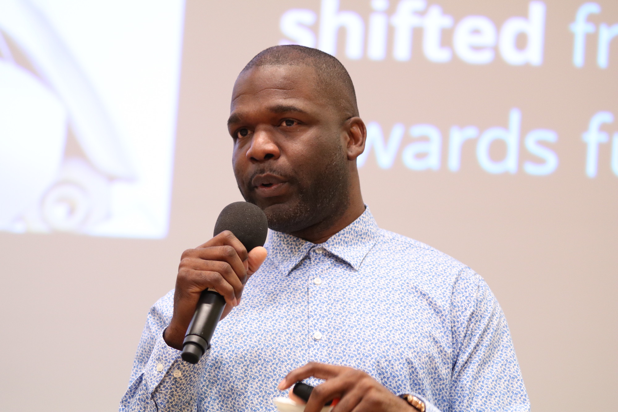 A close up photo of an African American male student speaking into a microphone, wearing a blue professional shirt