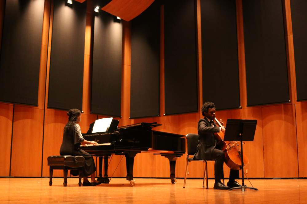 A Black female student plays the piano in a black shimmery dress while a Black male student wearing a tuxedo plays the cello