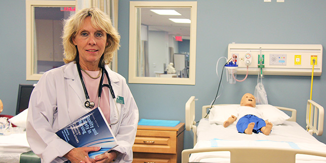 Dr. Teri Chenot posing with a textbook in front of one of the hospital beds in the STAR center with a child maniken