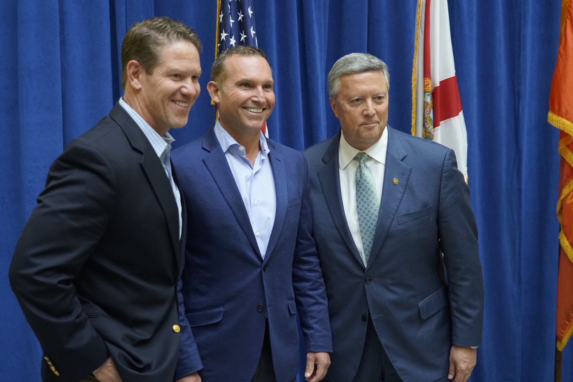 Mayor Lenny Curry and JU President Tim Cost, and another man are all dressed in blue in front of a podium, in front of a series of flags.