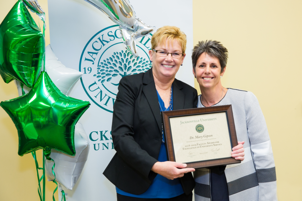 Dr. Mary Gipson and Dr. Christine Sapienza pose and smile for a photo while Dr. Gipson holds up her award plaque in front of a JU banner and green star balloons