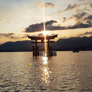 Photo of the Great Torii at sunset with mountains behind the floating gate and the sun shining right above the top of the shrine