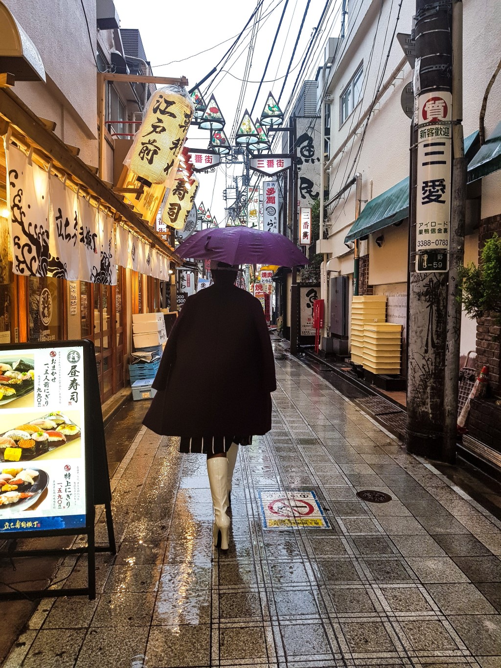 Student in boots and jacket with umbrella walking down rainy Nakano Broadway with lanterns overhead and stores nearby
