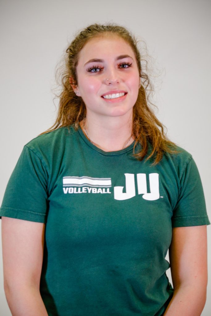 A female student smiling in front of a white wall, wearing a green shirt with a JU Volleyball logo on the front.