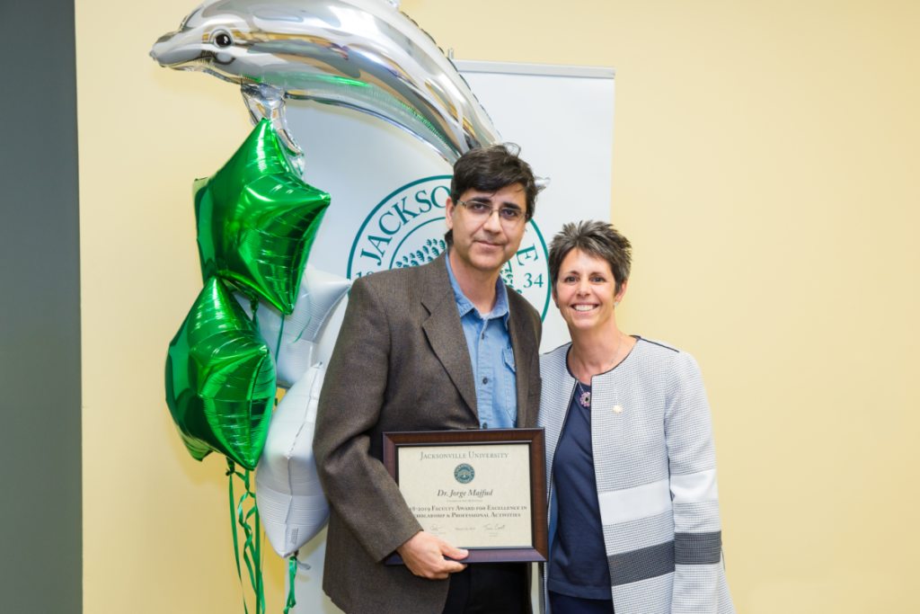 Dr. Jorge Majfud and Dr. Christine Sapienza pose and smile for a photo while Dr. Majfud holds up his award plaque in front of a JU banner and green star balloons