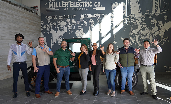 8 Miller Electric employees posing in front of an old car while holding their hands in the air in the JU hand sign
