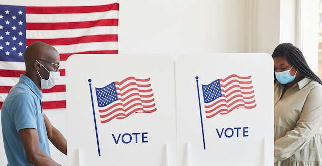 Photo of two people at voting stations with masks on placing their vote for an election with American flags on the voting booth and behind them