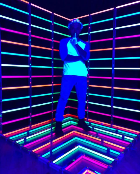 A male student standing in a dark room lined with neon lights.