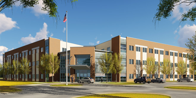 picture of a new building with tan and cream brick, three floors, and a number of windows. A cement roundabout is pictured at the bottom of the photo, and the American flag is at the top of the photo against a bright blue sky and a few clouds. 