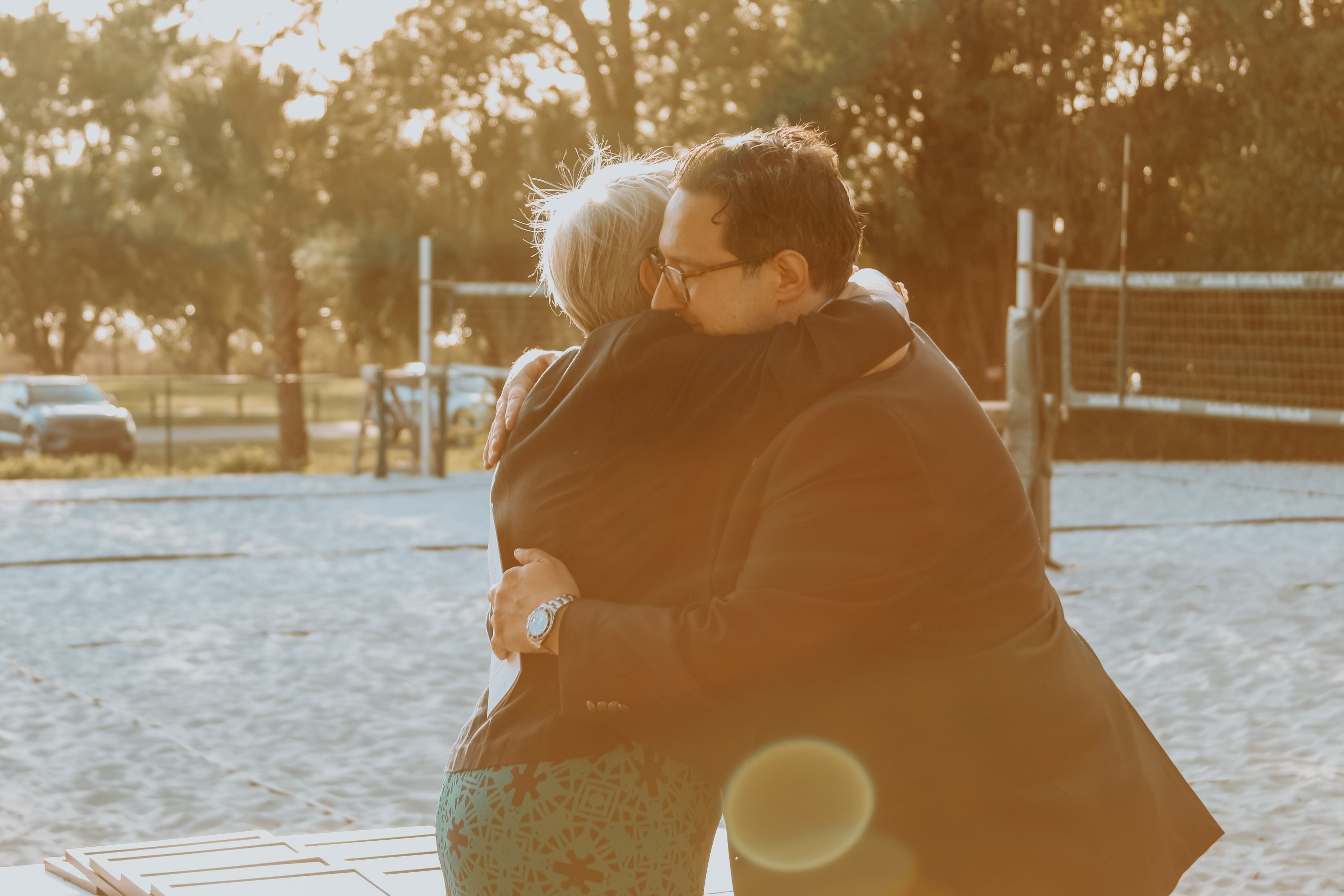 two middle-aged adults, one grey-haired female and one dark-haired male, hug at the conferring of an honor against the backdrop of trees and a river sunset.