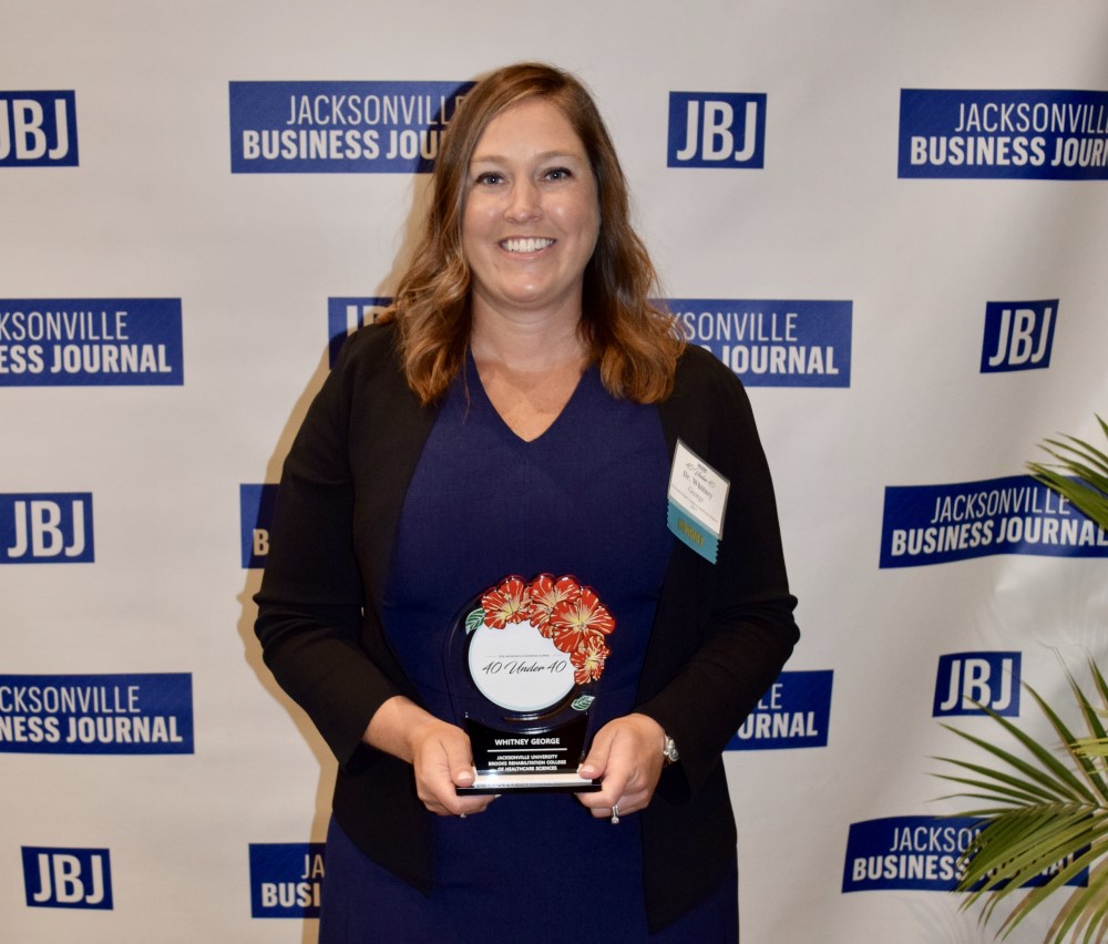 Dr. Whitney George posing with a 40 under 40 award infront of the Jacksonville Business Journal logo at the 40 under 40 event