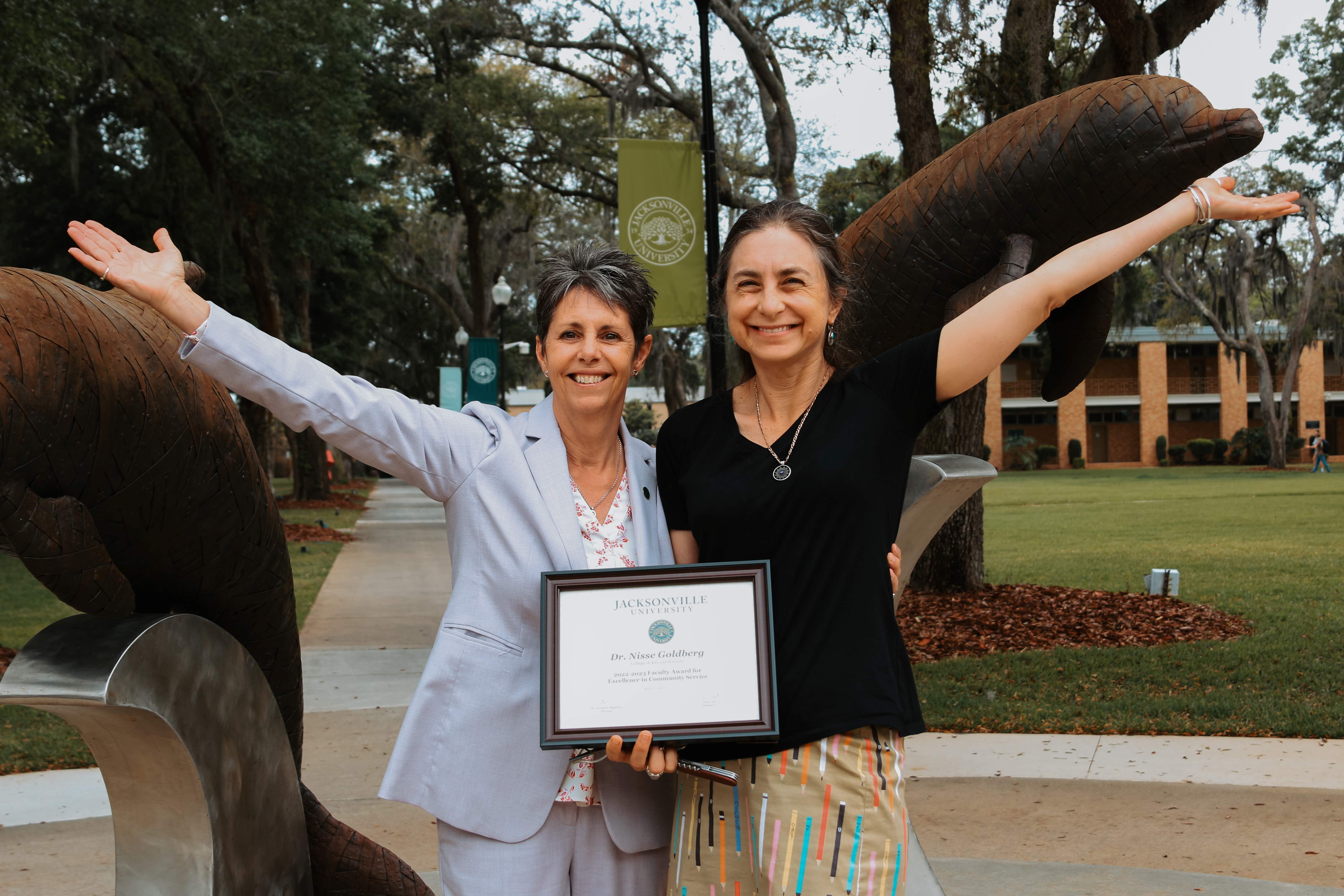 two women standing outside, one is holding an award