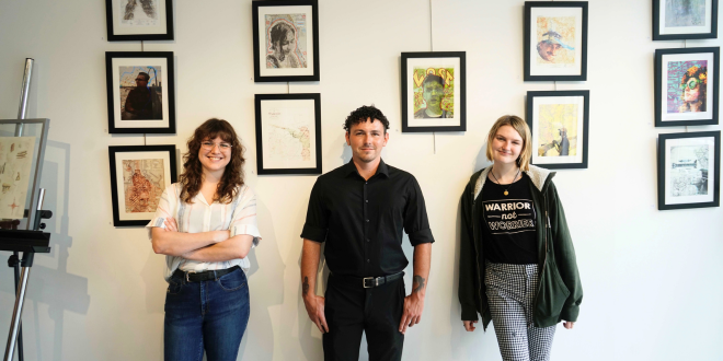 three students, two females and one male, are smiling against a backdrop of their artwork which includes colorful maps