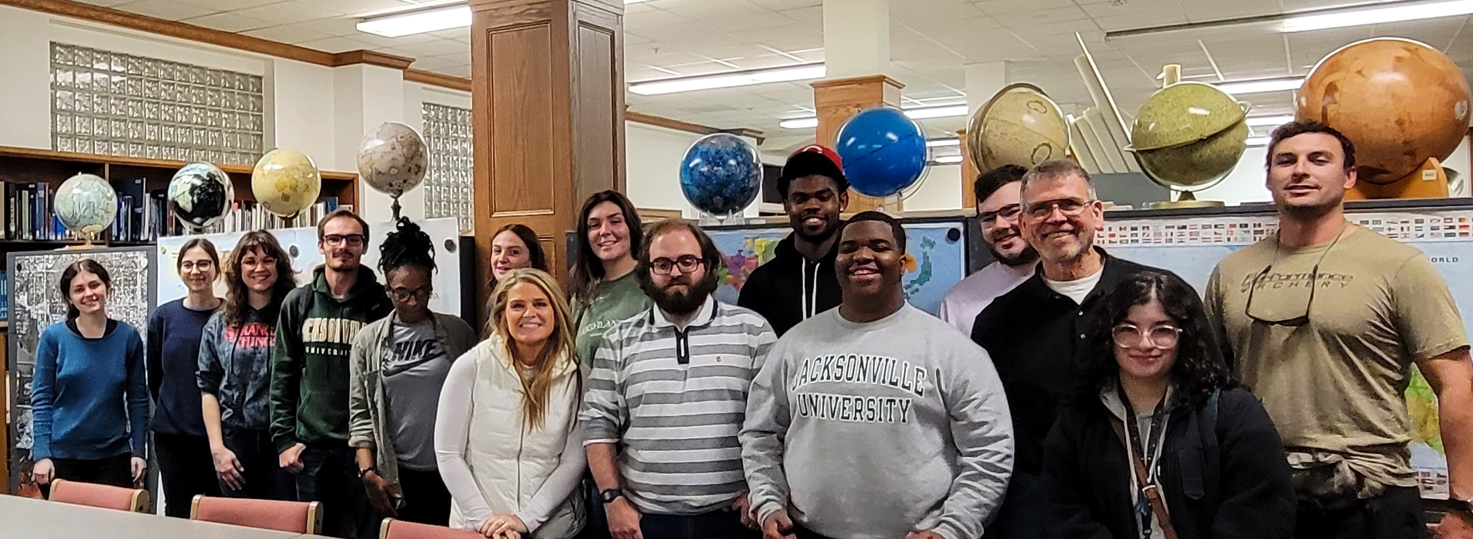 a large class of students and faculty stand smiling next to one another in a room filled with maps, globes, and posters