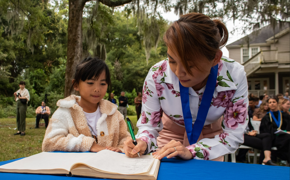 woman signing a book with a Phi Kappa Phi medallion on her neck and her daughter standing beside her