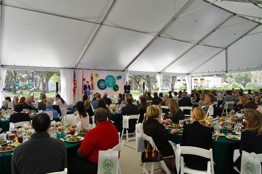 A large crowd seated at round tables under a white tent to listen to President Cost and Dr. Farrugia speak.