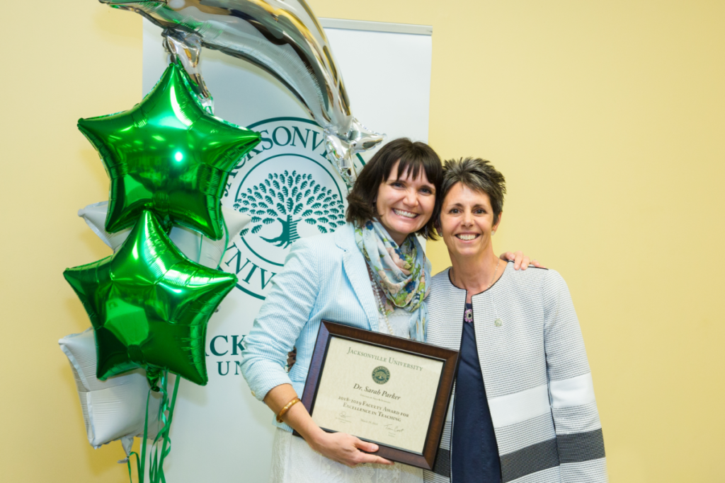 Dr. Sarah Parker and Dr. Christine Sapienza pose and smile for a photo while Dr. Parker holds up her award plaque in front of a JU banner and green star balloons